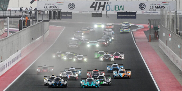 f1 Auto da corsa che competono nellAsian Le Mans Series allevento 4 Hours of Dubai. Le auto sono allineate in pista, sollevando polvere, con spettatori sullo sfondo e un grande striscione in alto.